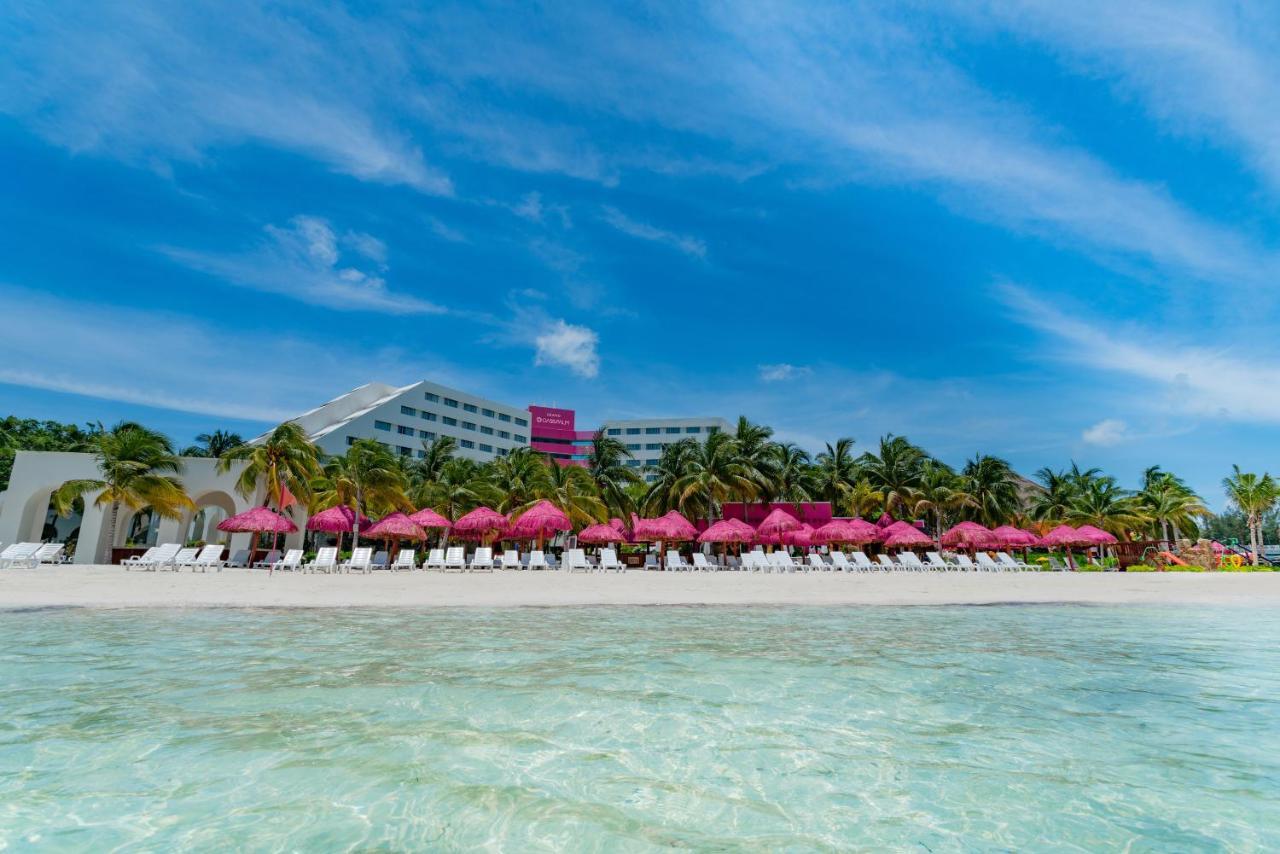 Grand Oasis Palm Hotel Cancun Exterior photo The photo depicts a tropical beach scene. In the foreground, there is clear, shallow water with a sandy bottom. In the background, a resort building is visible, with a row of pink umbrellas and lounge chairs set up along the beach. Palm trees surroun